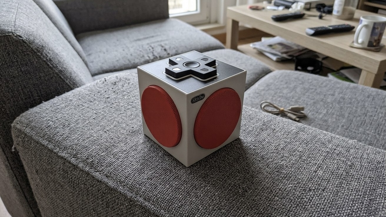 Колонка 8BitDo Retro Cube 2 в виде белого кубика с красными динамиками и крестовиной сверху стоит на подлокотнике дивана