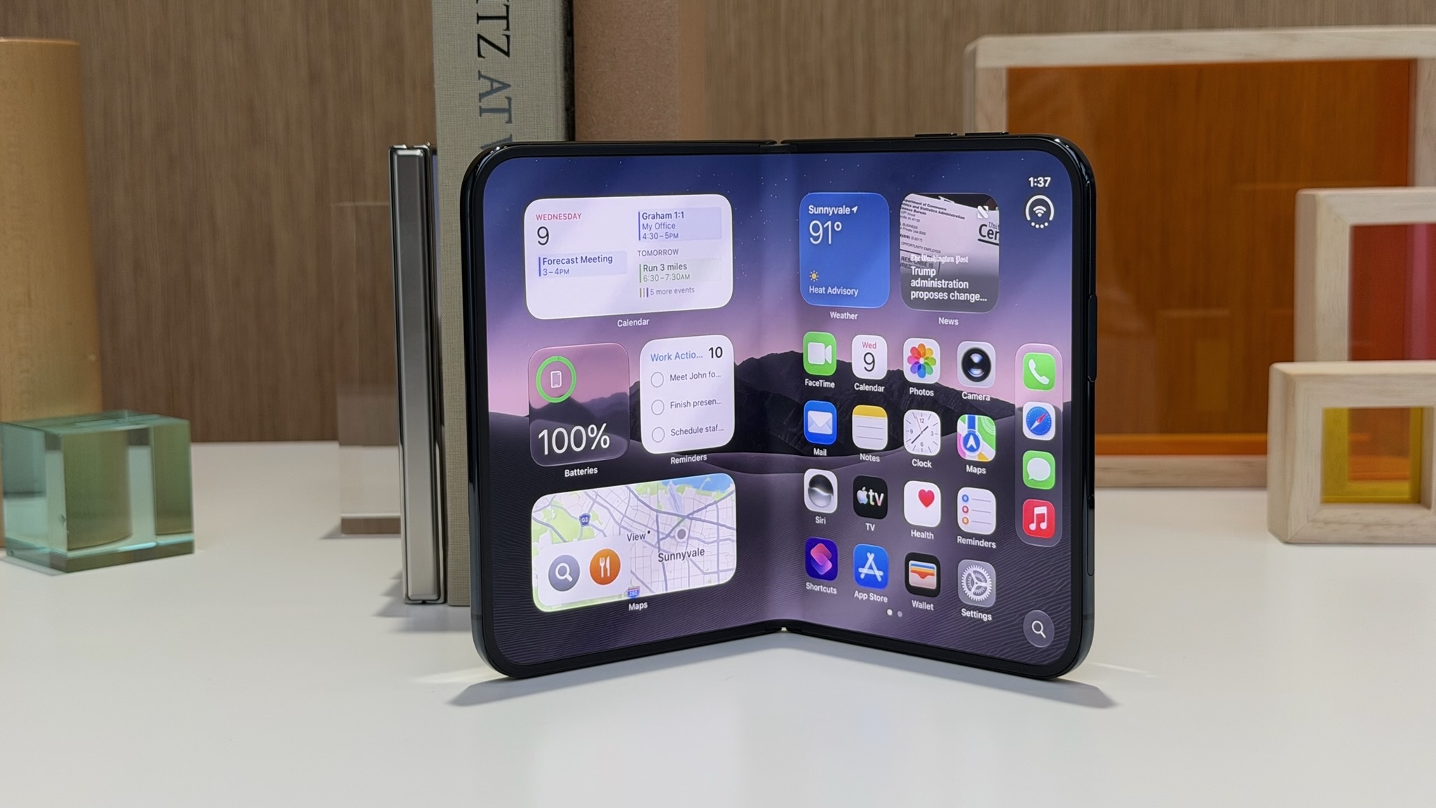 iPhone Duo стоит на столе в полусложенном виде, на внутреннем экране рабочий стол с виджетами