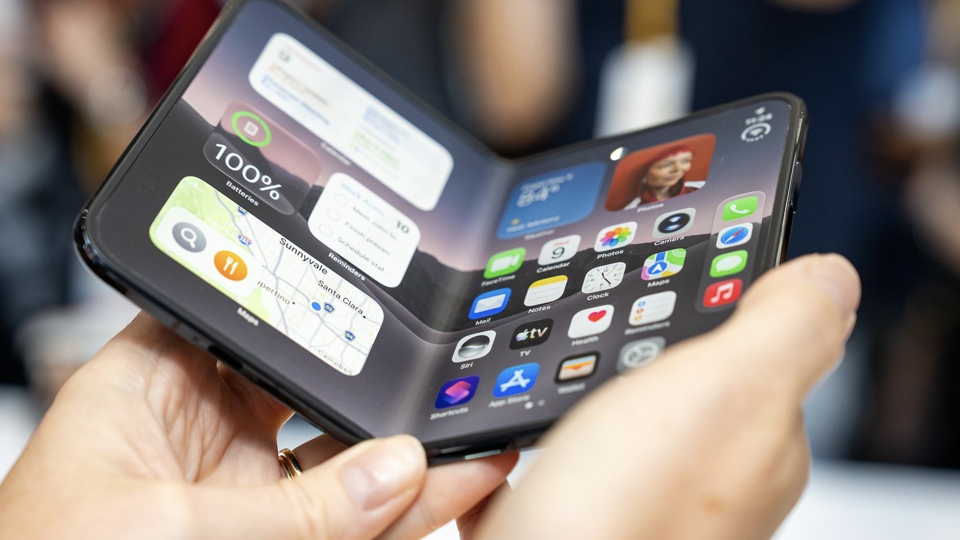 Полусогнутый темный iPhone Duo с виджетами Карт и Напоминаний в руках у человека