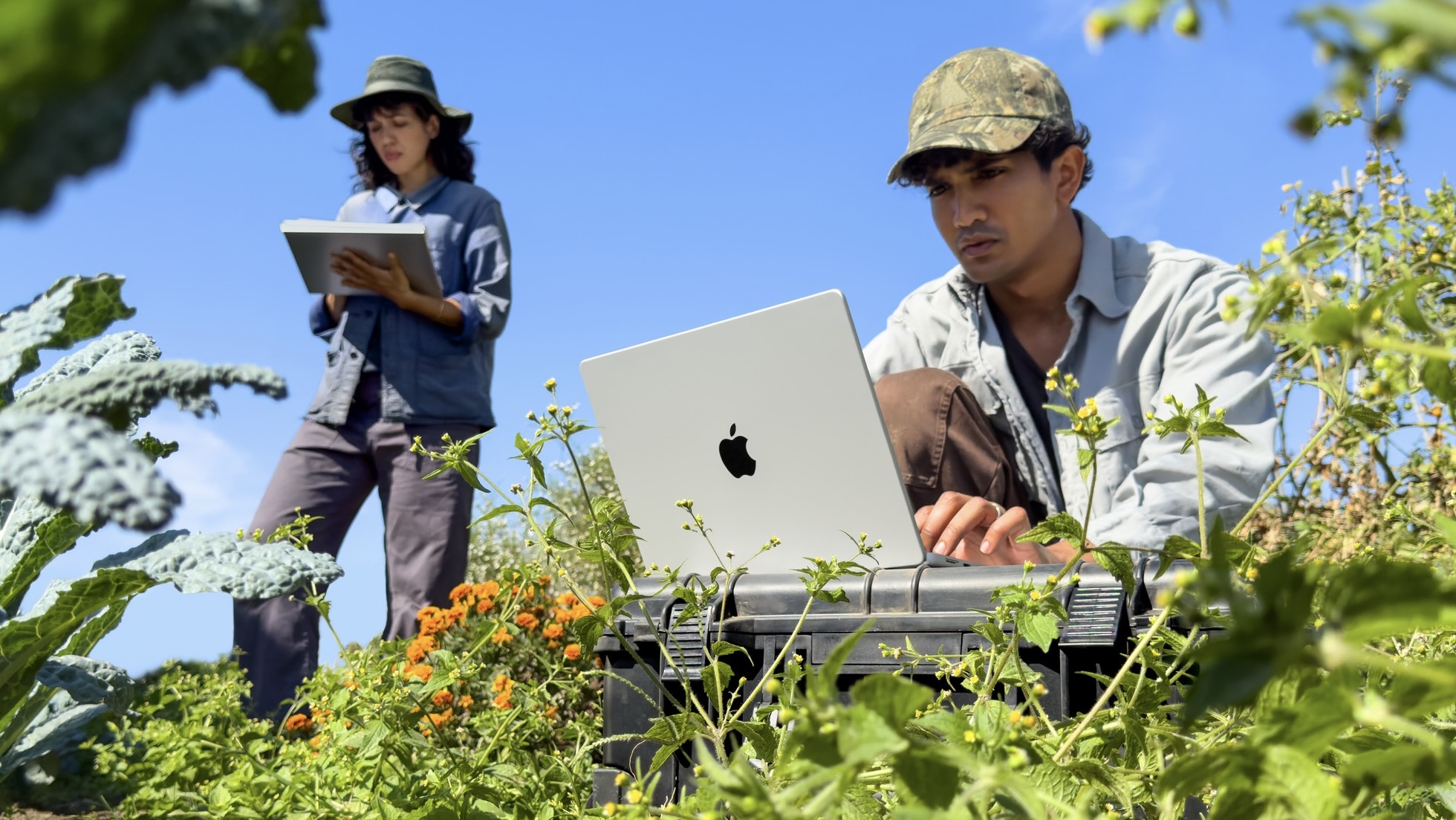 Мужчина работает на серебристом MacBook Pro в огороде, рядом девушка держит iPad