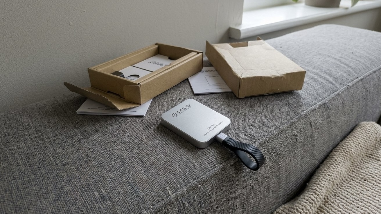 Распакованный SSD ORICO K15 Mini с кабелем-петлей лежит на сером диване рядом с коробкой