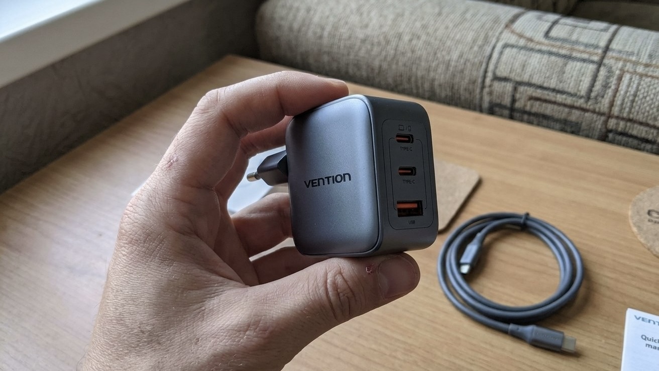 Серый адаптер Vention на 70 Вт с двумя портами USB-C и одним USB-A в руке, на столе рядом лежит кабель USB-C